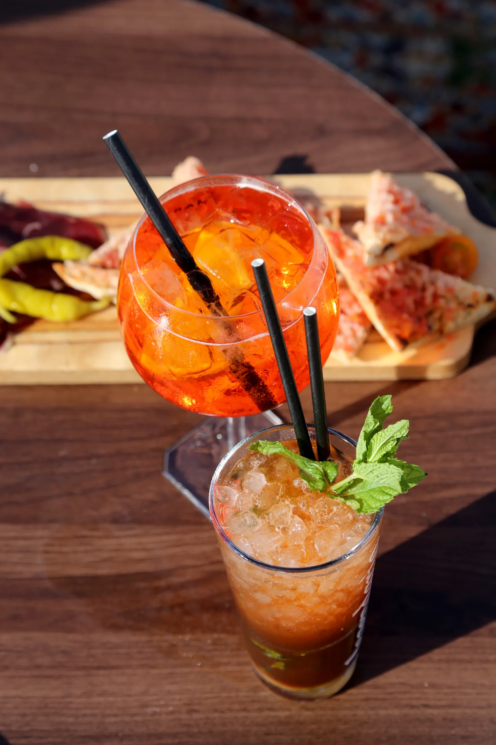 Two cocktails on a wooden table with snacks in the background.